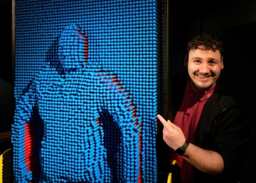 A male tour guide stands beside a Pinwall exhibit, a wall of plastic blue pins