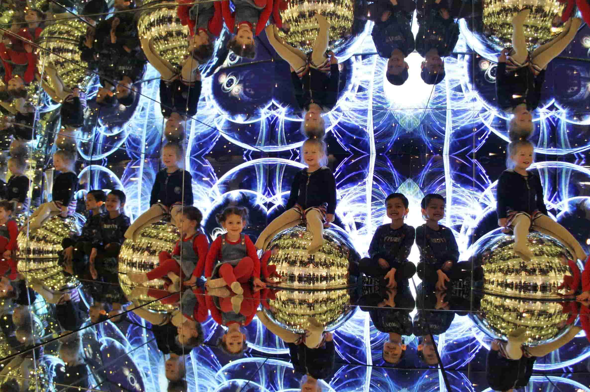 Children sit in a large mirrored box with colourful patterns in the background